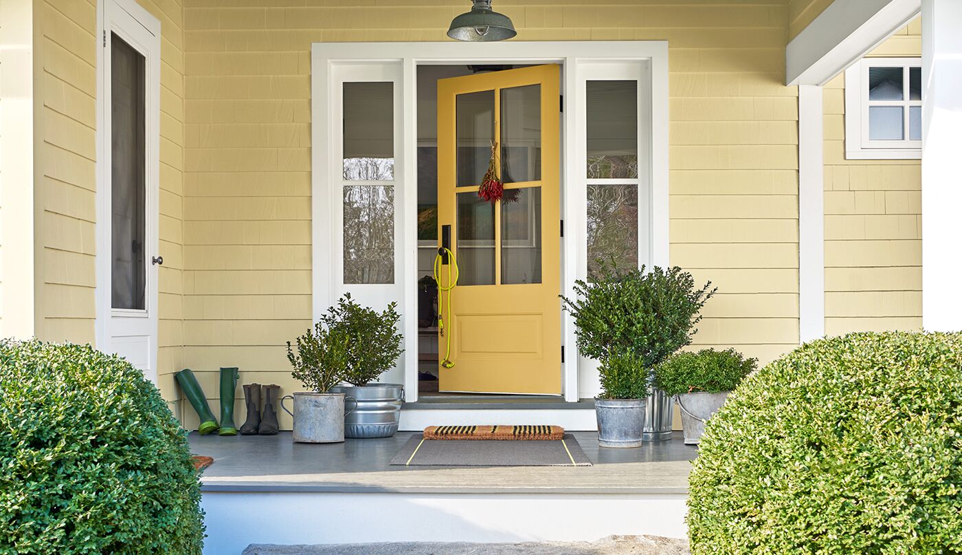 Two green potted plants sit next to a yellow door on a porch of a yellow siding house.