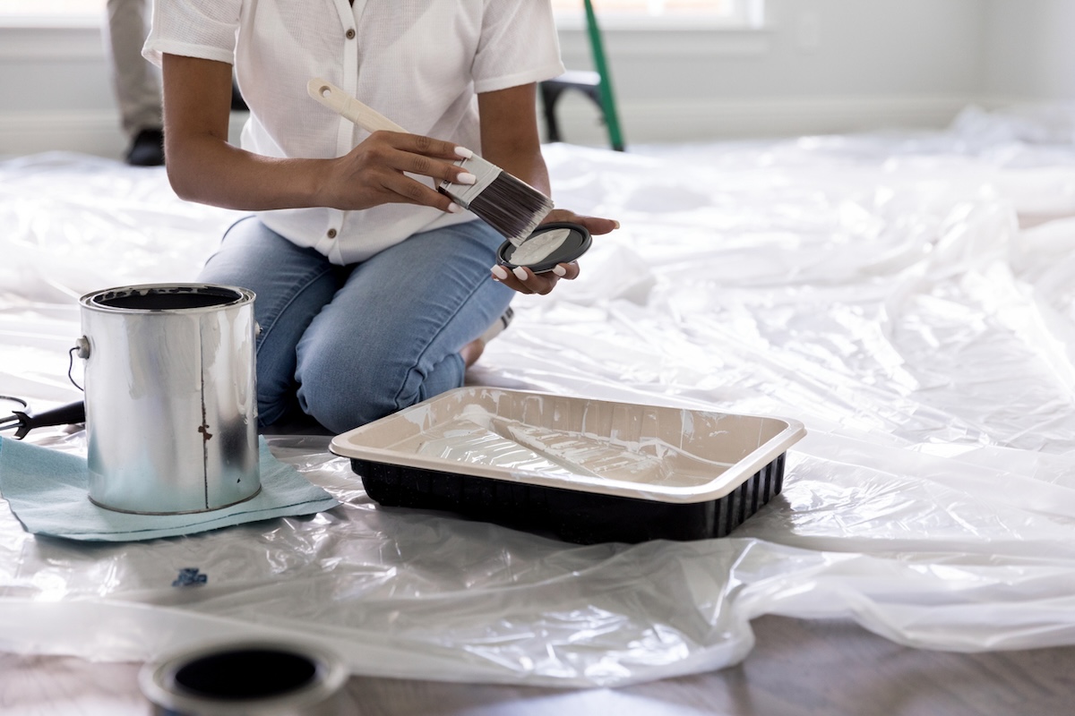 Woman kneeling on the floor of a living room holding a paint brush and preparing to paint the walls.