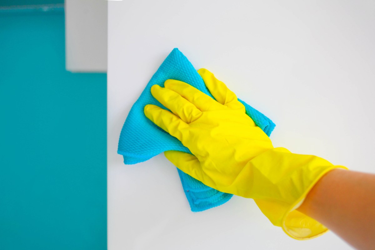 A female hand in yellow rubber gloves wipes a modern white kitchen wall with a microfiber cloth.