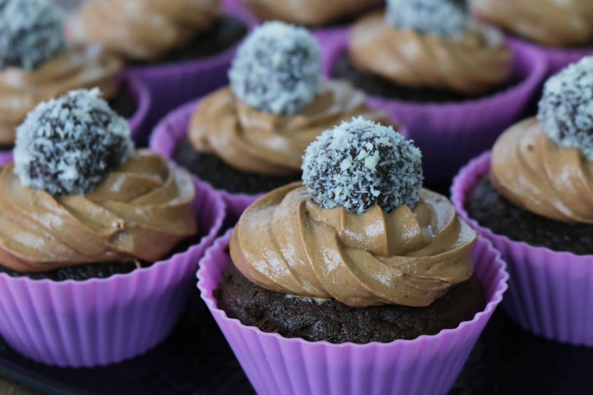 Close view of tray of homemade chocolate cupcakes topped with piped chocolate icing frosting and coconut covered truffles in purple silicone cake cases.