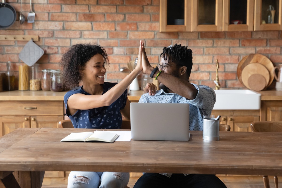 Excited young African American couple sit at desk at home kitchen give high five celebrate online lottery win on laptop. Happy biracial man and woman enjoy success working on computer. Luck concept.