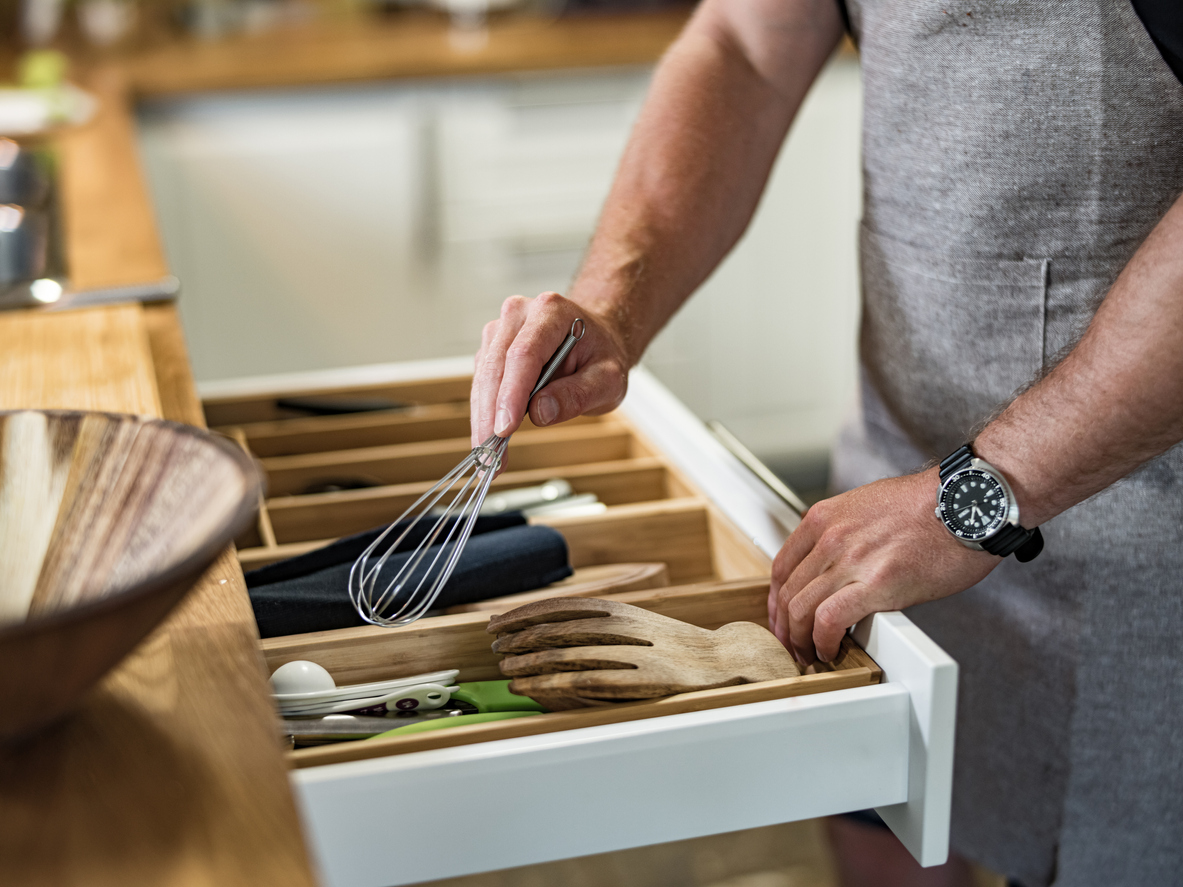 Hands of a man taking the wire whisk from the utensil drawer in a home kitchen. He is wearing casual clothes with chef's apron.