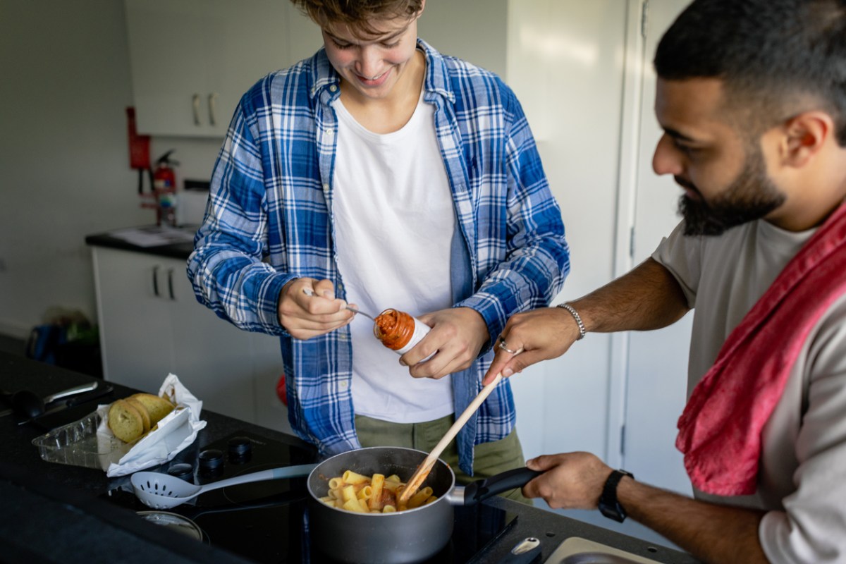 A front view shot of male uni student friends standing in their kitchen, they are wearing casual clothing and preparing a meal together. One man is stirring pasta in a cooking pan as they add the sauce.