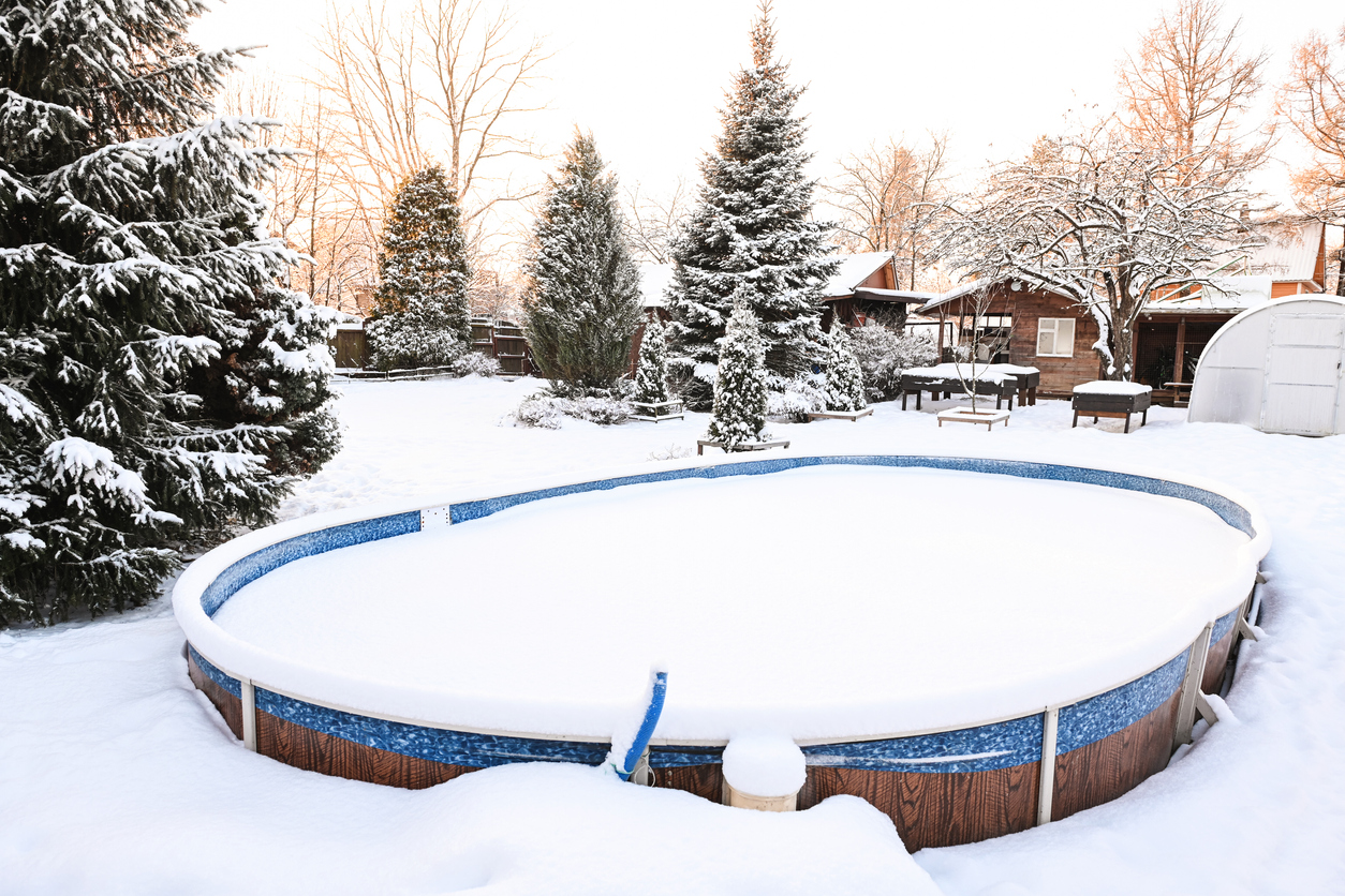 Snow-has-piled-onto-the-cover-of-an-above-ground-pool-on-a-home-property-among-snowy-evergreen-trees.