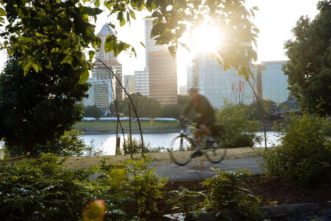 A cyclist rides on a bike path along the river in Portland with a view of the city across the water.