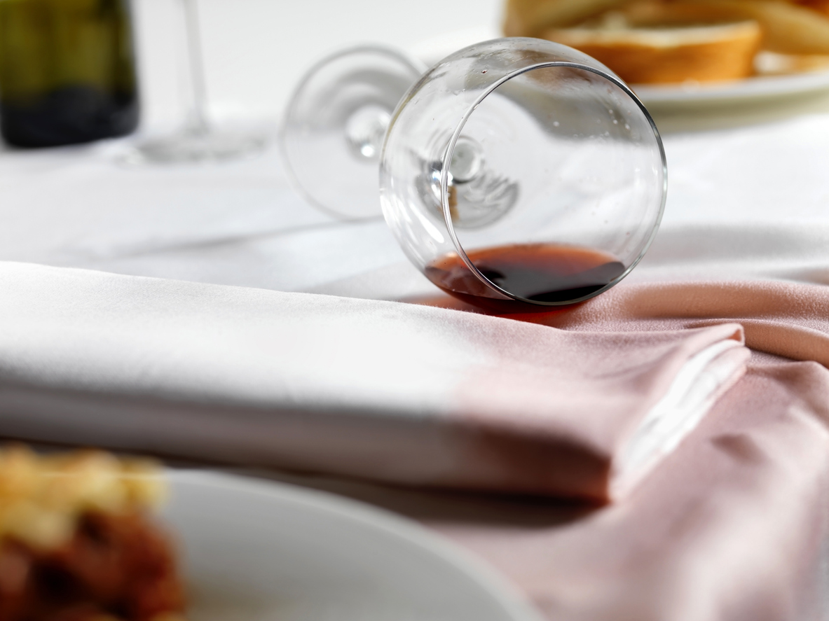 A place setting on a table with a white tablecloth and napkin with a glass of red wine spilled and staining the tablecloth.