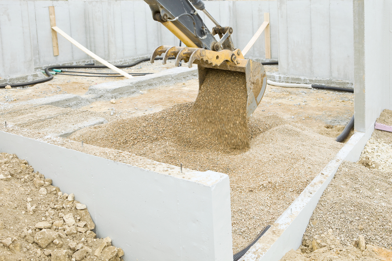 An excavator bucket is dumping rock inside a new house foundation wall. The rock is laid on top of the dirt and drain tile for a radon mitigation system. A concrete slab will be poured on the rock forming the floor.