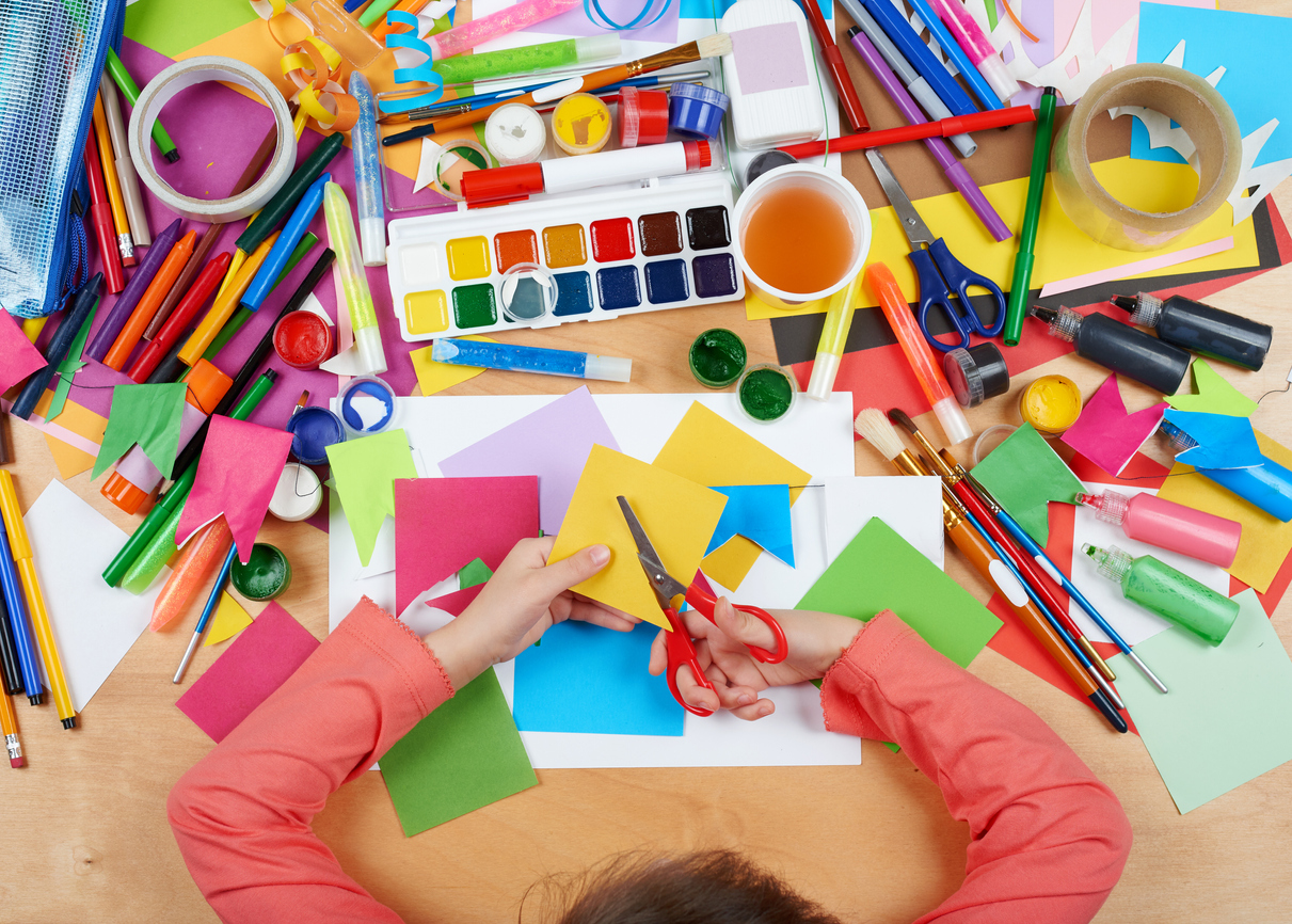 An overhead view of a child doing arts and crafts at a table covered in paper, paint, and other supplies.