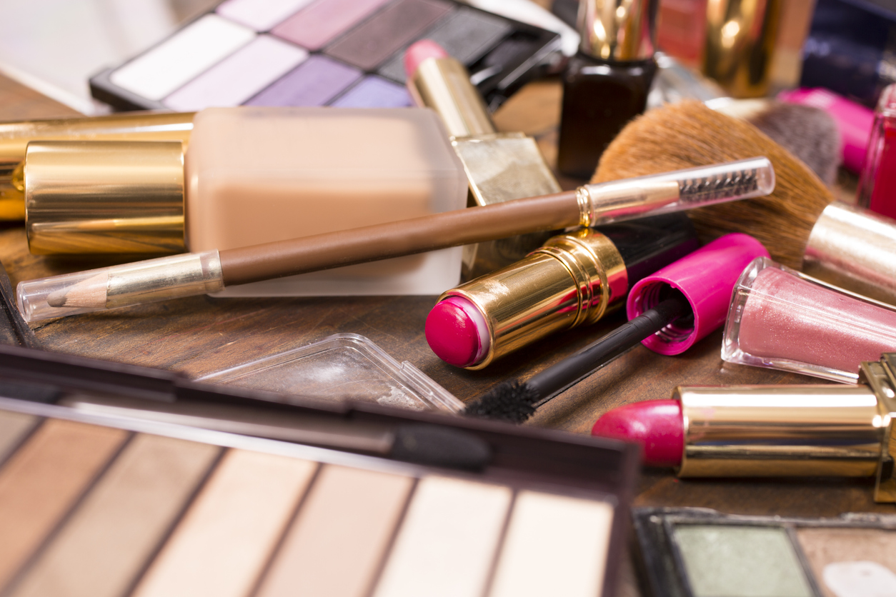 Large pile of various cosmetics on top of a wooden dressing table. Make-up items include: eye shadow, nail polish, foundation, lipstick, make-up brushes, blush, mascara, lip gloss.