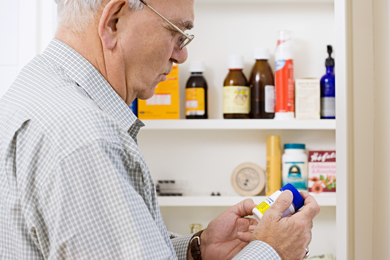 Man reading label on bottle of tablets in front of open medicine cabinet filled with pill bottles.