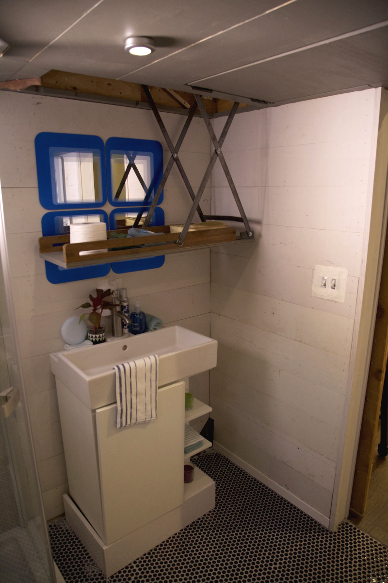 The corner of a tiny house bathroom with a pull-down shelf installed into a cutout in the ceiling above a sink with built-in shelves.
