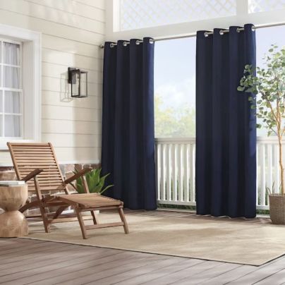 The Sunbrella Solid Outdoor Grommet Curtain hung along a front porch next to a tree and lounge chair.
