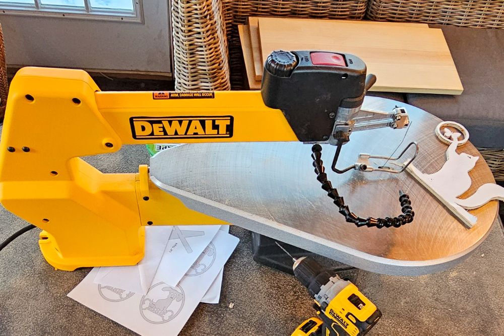 The DeWalt scroll saw on a workbench with a recently cut wooden decor item, paper stencils, slabs of uncut wood, and a DeWalt drill.