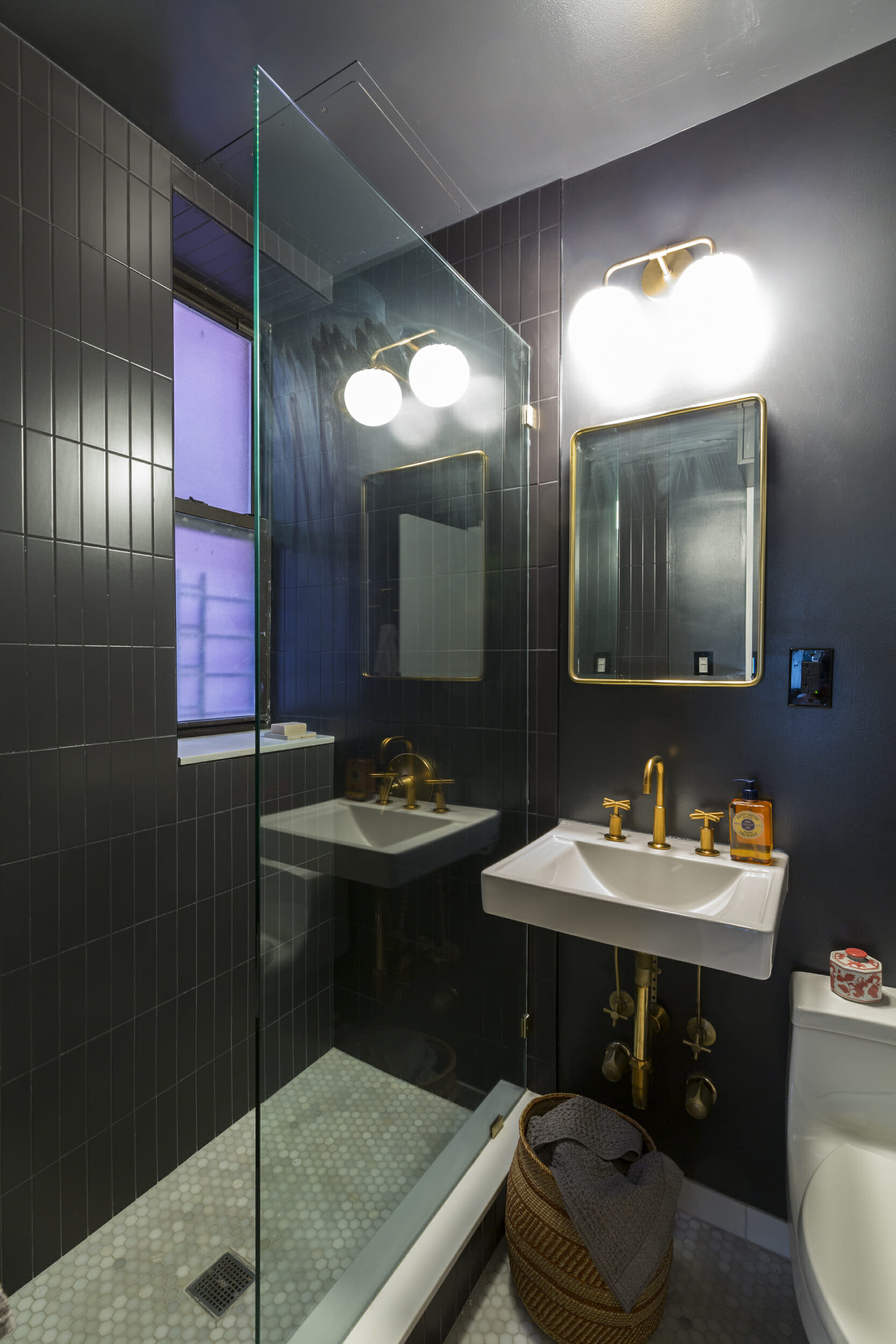 A tiny bathroom with black walls and gold sink, shower, and lighting fixtures.