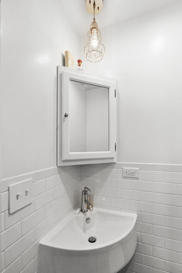 The bright corner of a white bathroom with a corner sink and medicine cabinet.