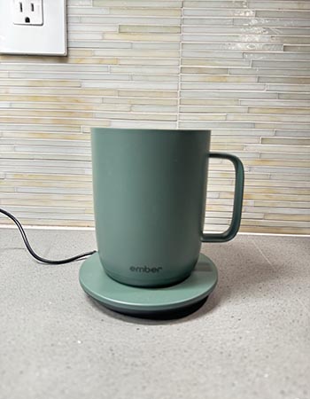 The Ember Mug 2 in sage green on a kitchen counter on its plugged-in charging coaster.