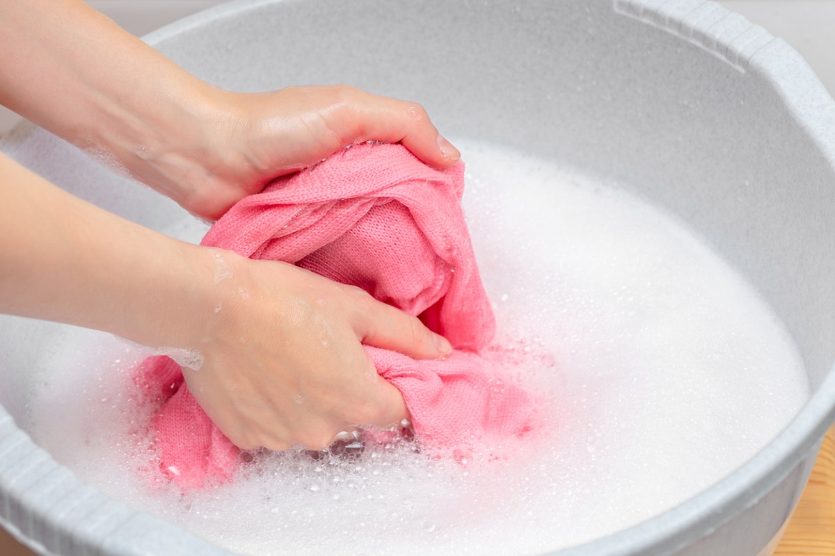A person hand-washing a pink sweater in a white basin full of soapy water.