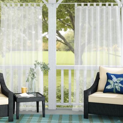 The Winston Porter Hottinger Solid Sheer Outdoor Curtain installed at the edge of a porch to provide privacy for a seating area.