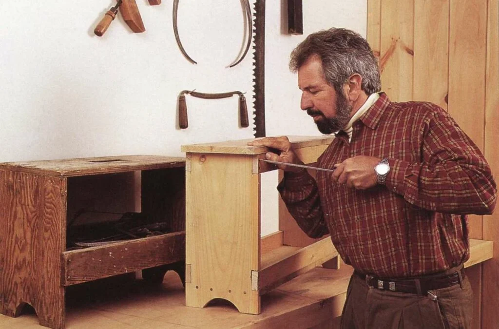 Bob Vila in a plaid shirt uses a screwdriver to work on a wooden bench.