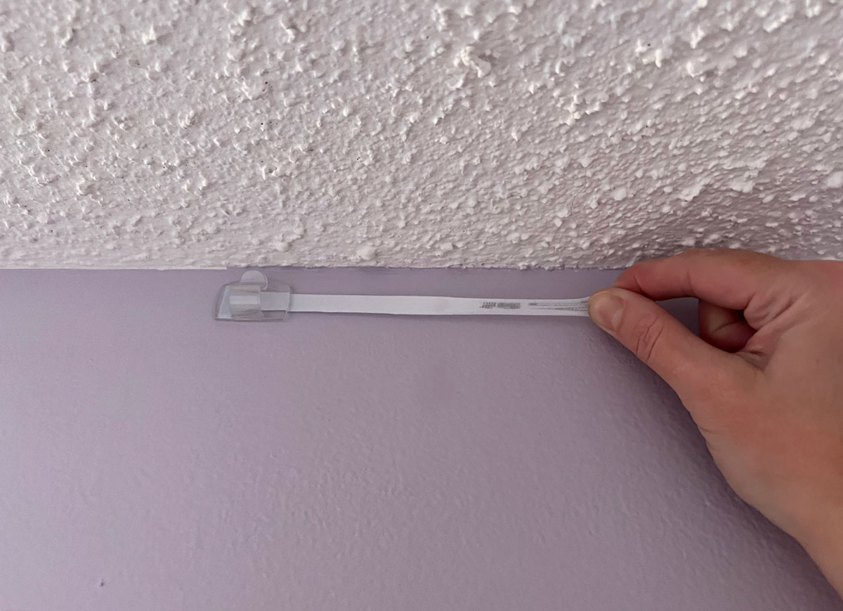 A person removing a 3m command strip stuck to the top of a wall inside a home.