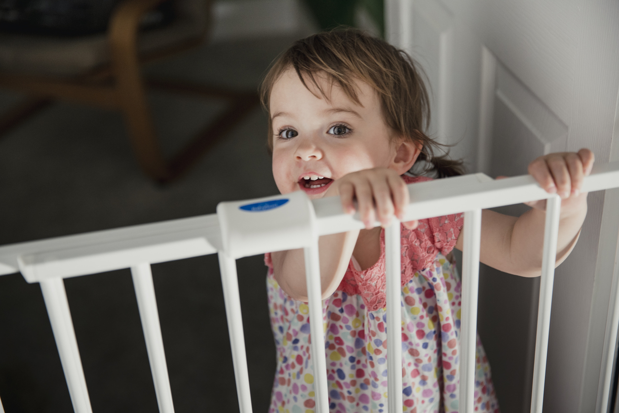 Front view of a little girl standing on the other side of a baby saftey gate. The little girl is holding onto the top of the saftey gate and looking at the camera over the top.