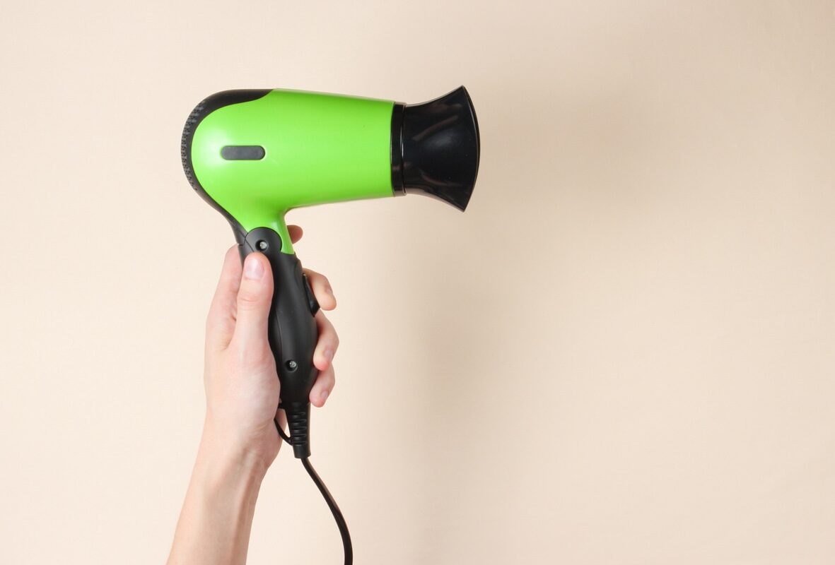 A hand holding a green hair dryer up to a beige wall.