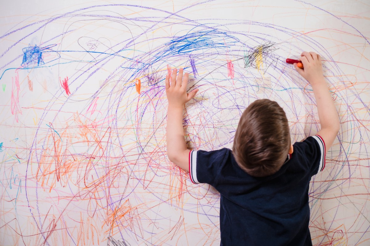 Little boy with brown hair and navy blue shirt colors white wall with crayon.