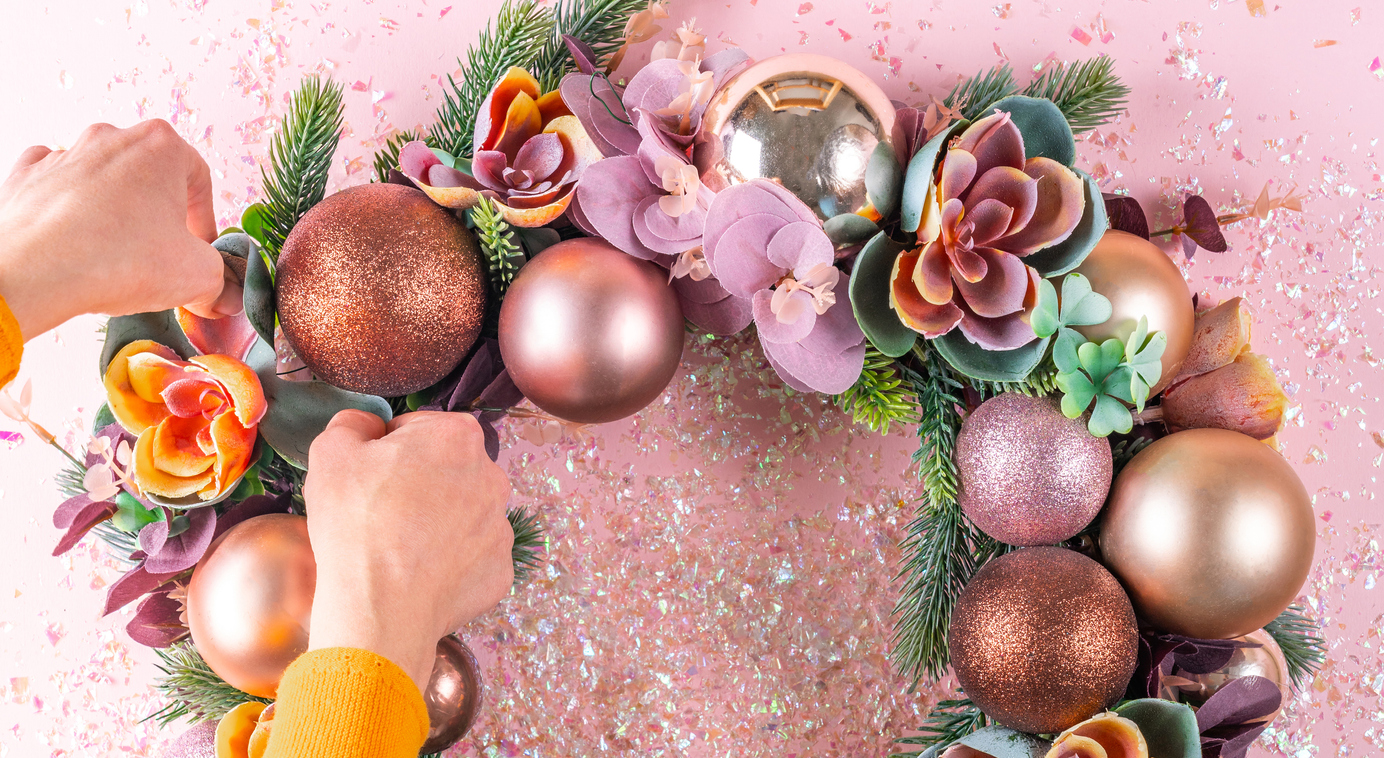 Hands-arrange-a-faux-flower-on-a-pink-wreath-with-greenery-and-Christmas-tree-ornaments.
