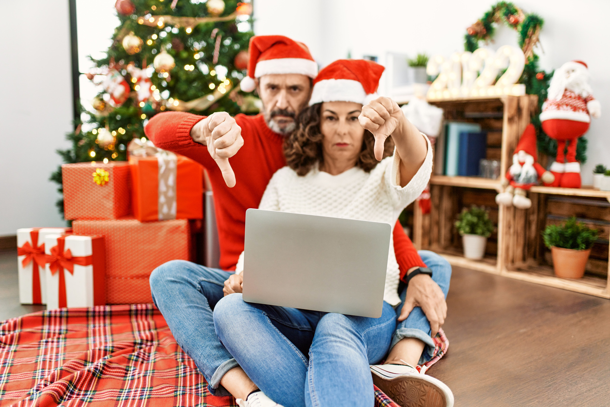 Hispanic middle age woman and mature man using laptop sitting by christmas tree with angry face, negative sign showing dislike with thumbs down, rejection concept