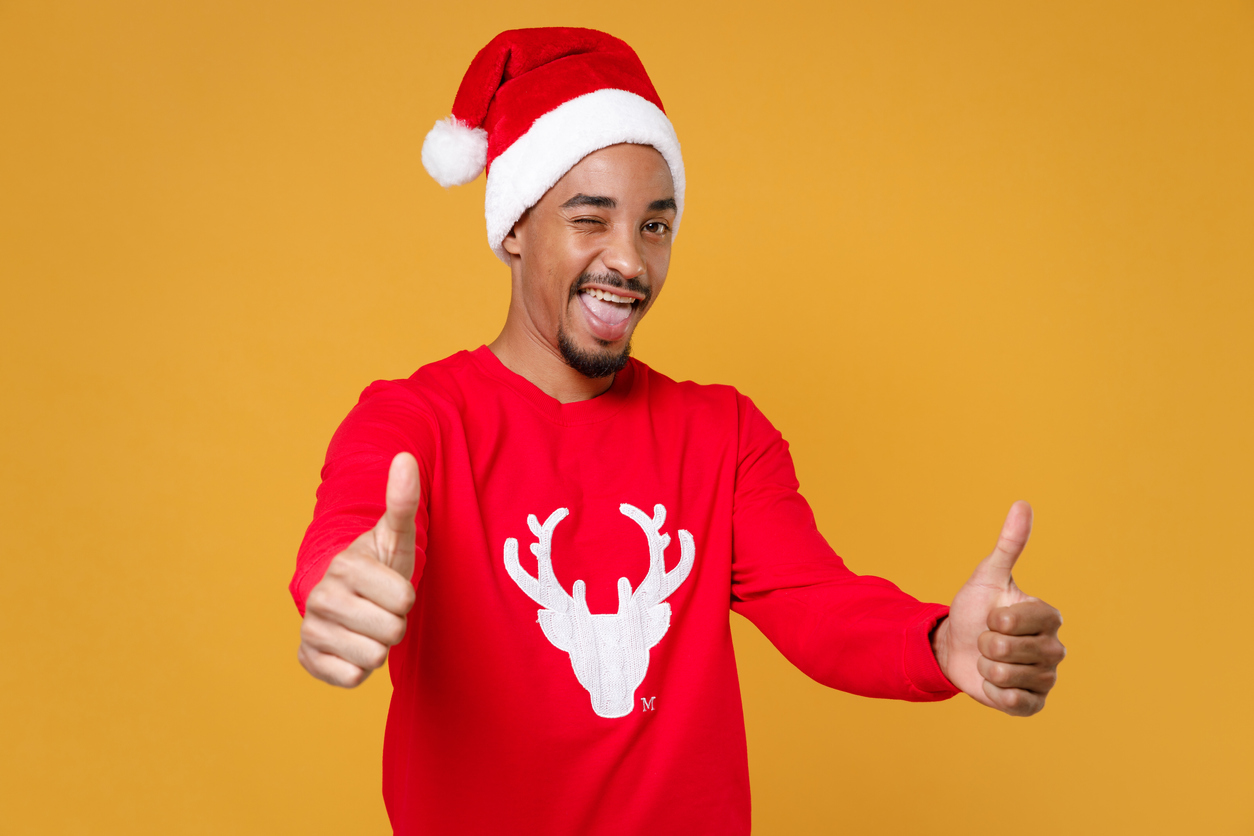 Blinking young Santa african american man 20s in red sweater with deer Christmas hat showing thumbs up isolated on yellow background studio portrait. Happy New Year celebration merry holiday concept.