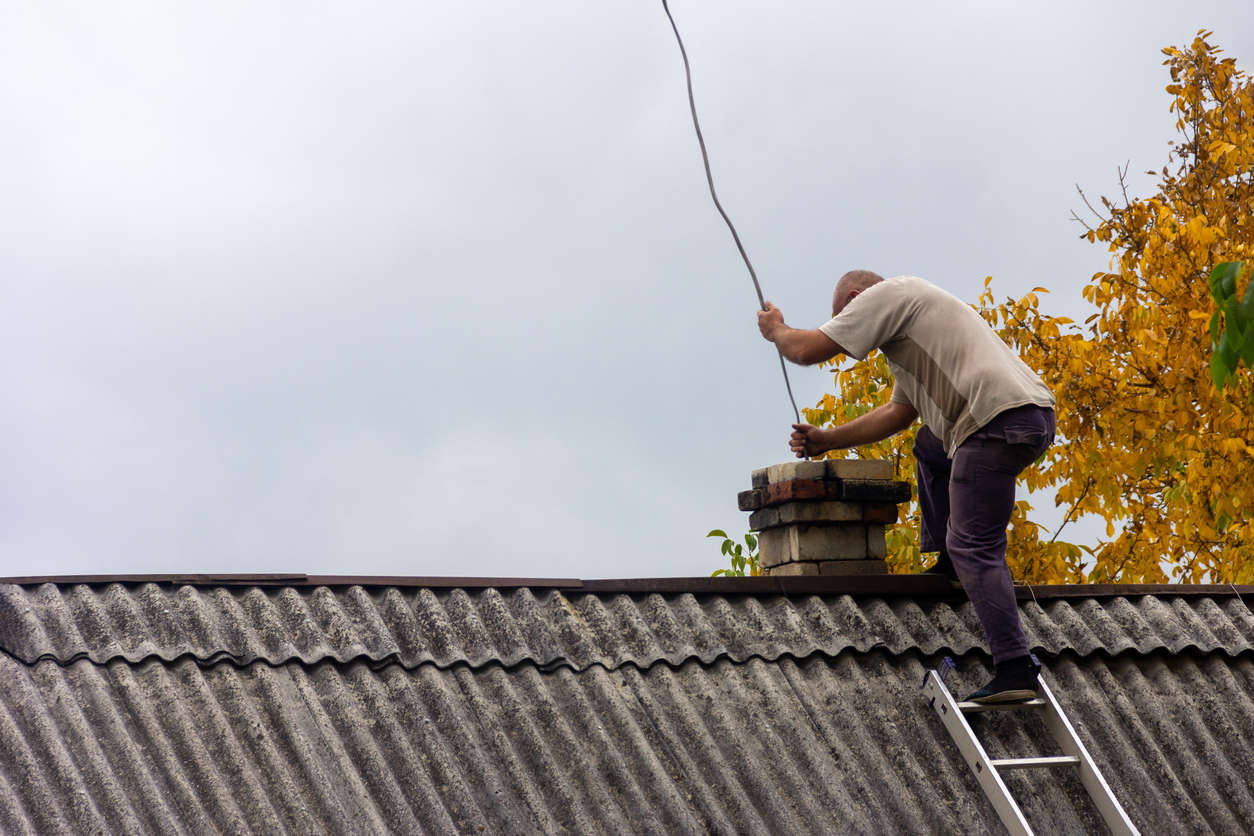 a chimney sweep man cleans the chimney from soot on the roof of a village house against a gray sky with a copy space, preparing for the heating season