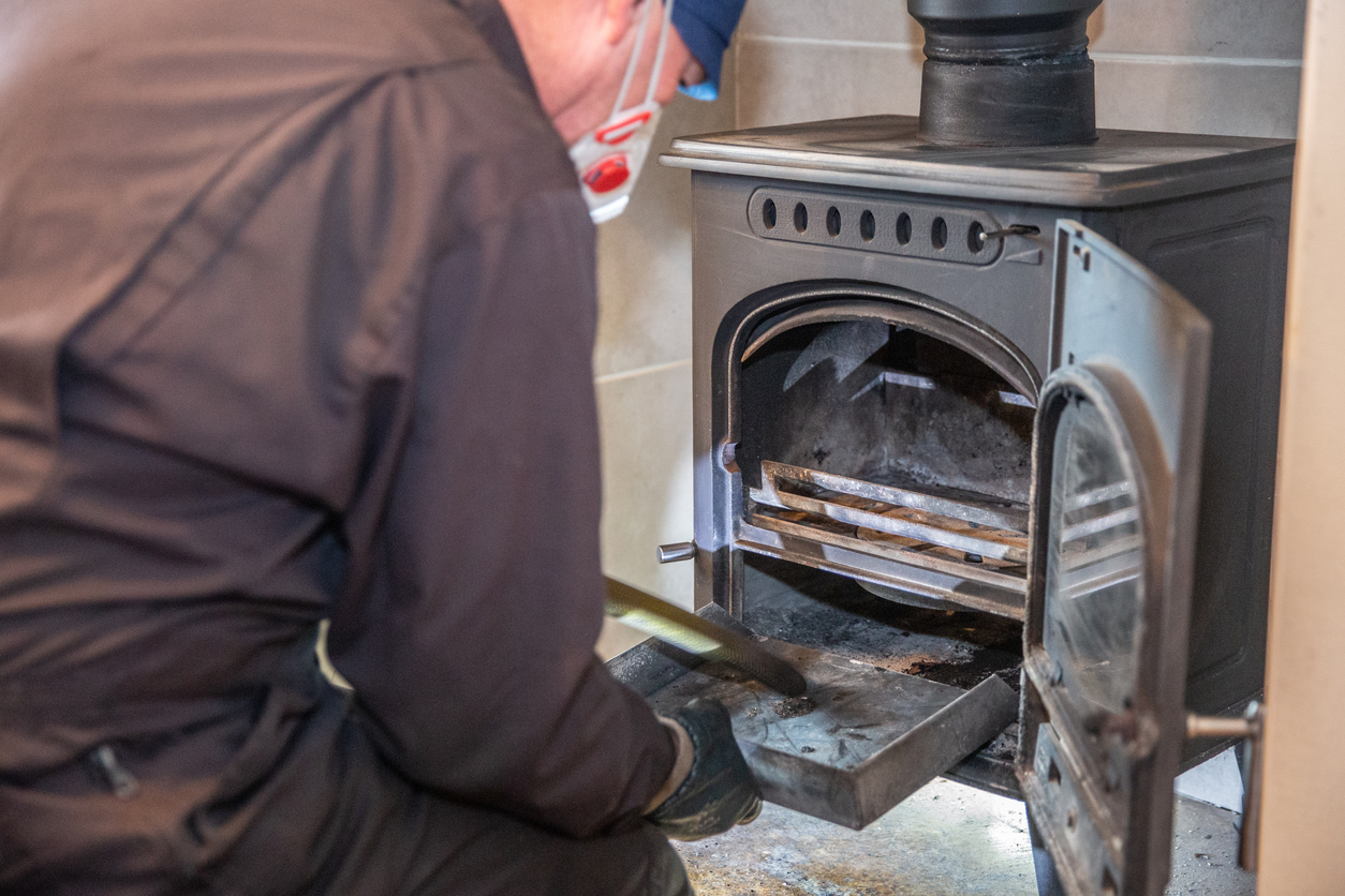 A-man-in-coveralls-and-a-face-mask-cleans-ashes-from-a-wood-burning-stove.