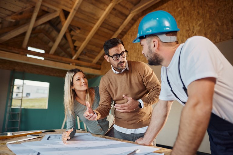 Young couple talking to manual worker about housing design plans at construction site.