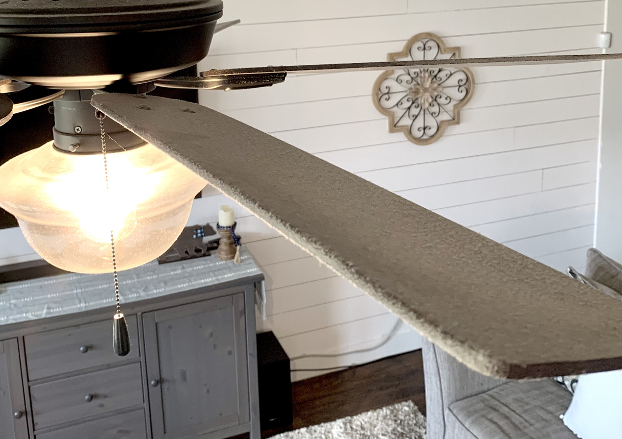 A-ceiling-fan-blade-over-a-grey-and-beige-living-area-is-covered-with-dust.