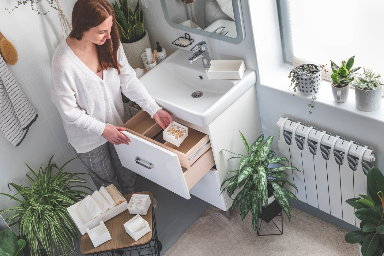 Happy young woman standing in front of open drawers of dressing table and taking out toiletries from drawer. Concept of saving space and organizing vanity table. Houseplants.