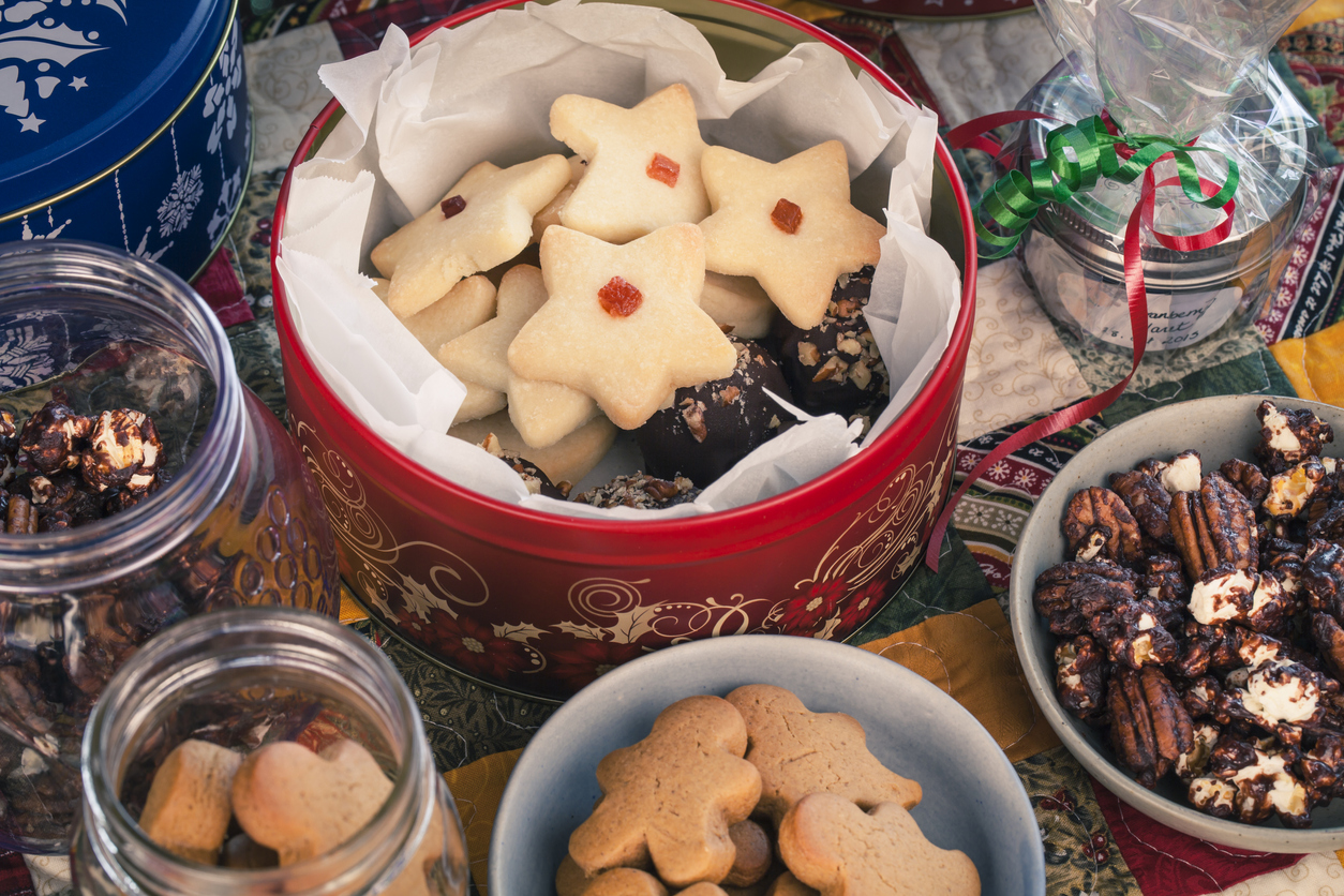 Colorful bird's eye view of various shortbread and gingerbread cookies in tins and chocolate popcorn and cranberry claret wrapped in cellophane all on a patchwork quilted table runner.