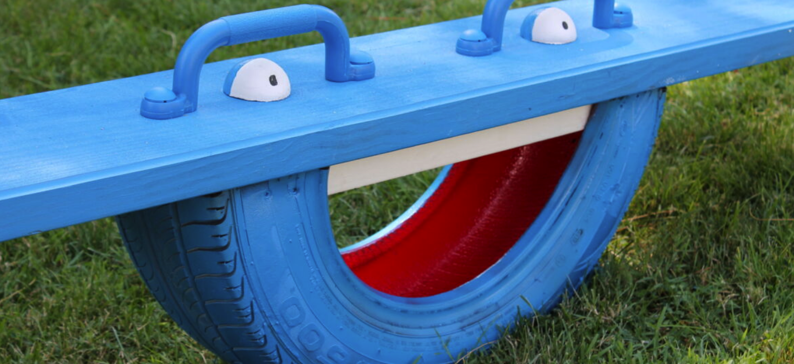Close view of a seesaw made from an old tire painted blue with a cartoon face and blue plank on top