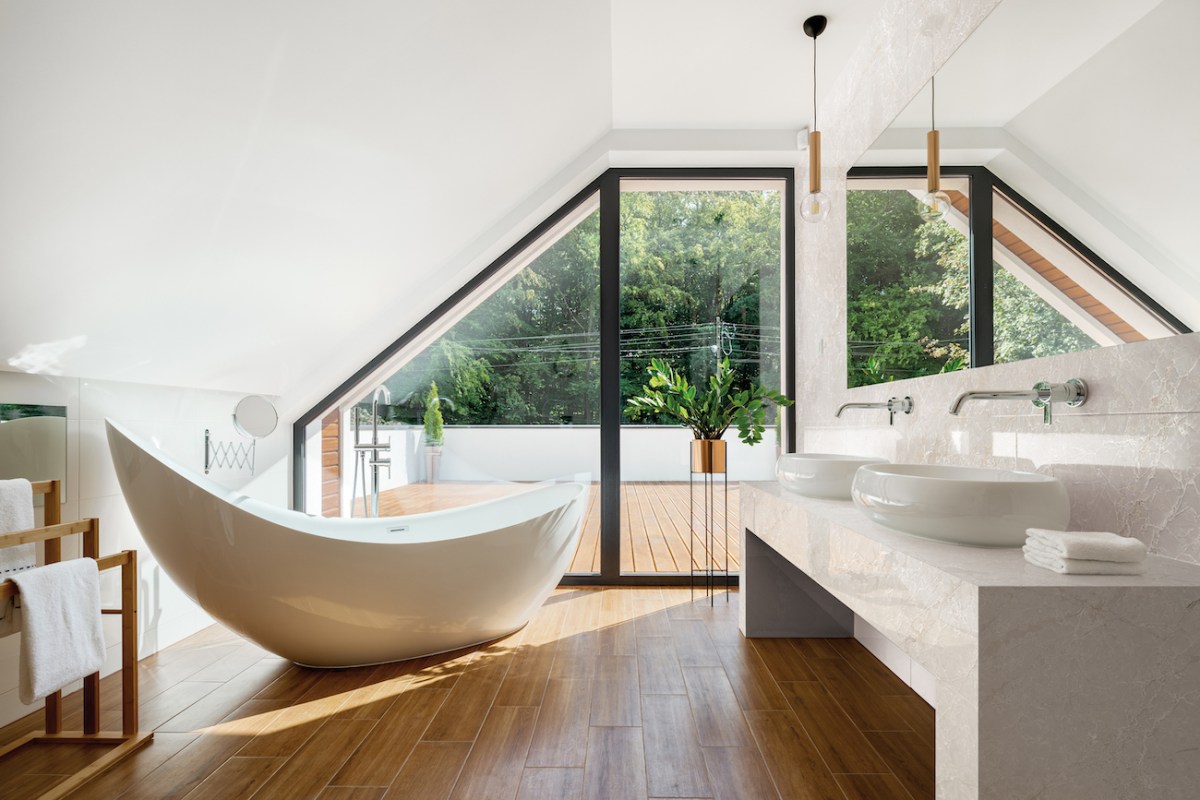 An airy white bathroom has wood floors and a free standing curved tub in front of a large window.