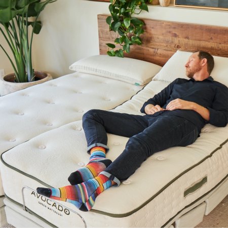  A person reclining on the Avocado Eco Pro Adjustable Base that's installed against a wood headboard in a room with multiple plants.