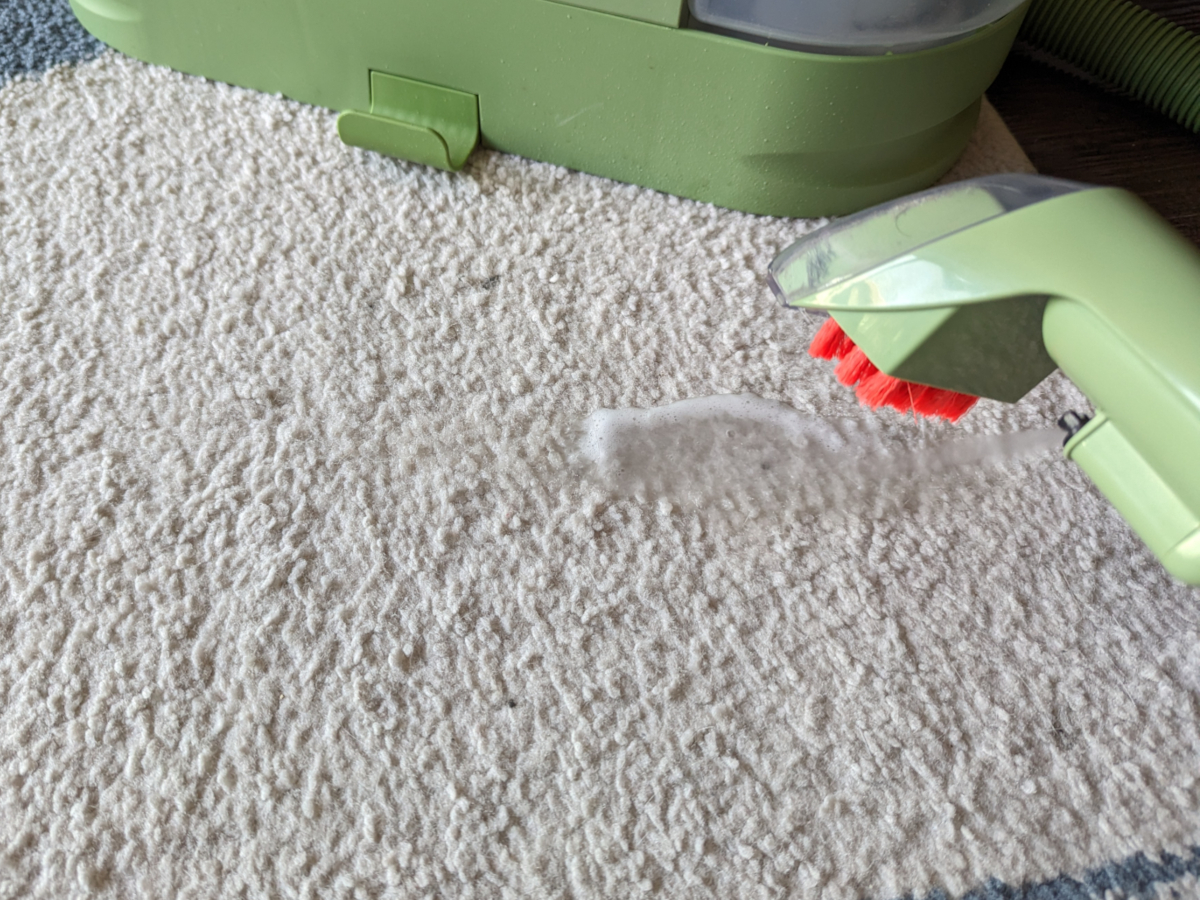 An area rug stain being sprayed with soapy water solution by a Bissell Little Green cleaning attachment.