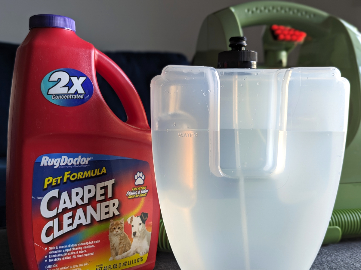 A Bissell Little Green carpet cleaner's filled water tank in front of the appliance and a bottle of pet carpet cleaner formula.