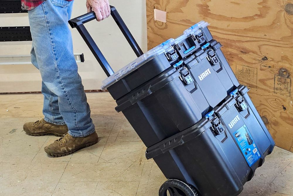 A person holding the telescoping handle of the Hart Tools stack system tool box while wheeling it through a workroom.