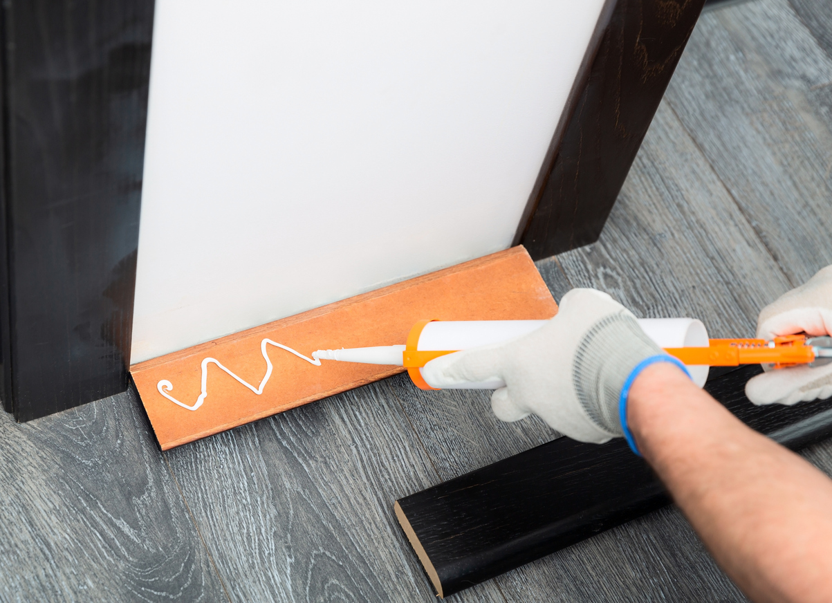 Handyman is putting a caulk or glue from tube on a wooden baseboard before attaching it to the wall.