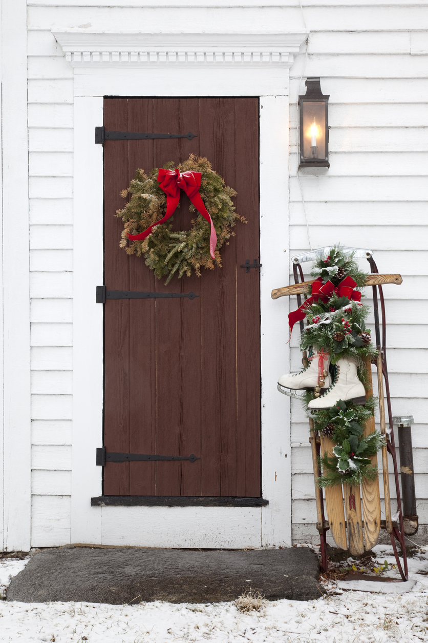 Antique sled and skates propped up against white home siding, with Christmas wreath on adjacent brown front door.