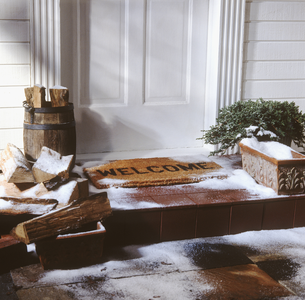 Front porch in winter, with a doormat, firewood in barrels, and other decor.