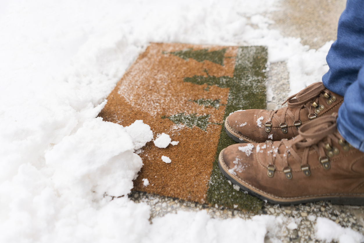 A man's winter boots standing outside on a welcome mat in the snow.