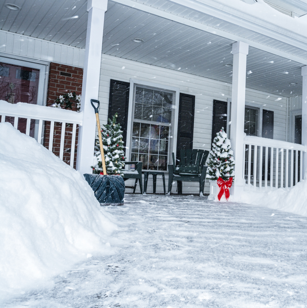A front porch covered in snow with a snow shovel propped against the railing and patio furniture covered in snow.