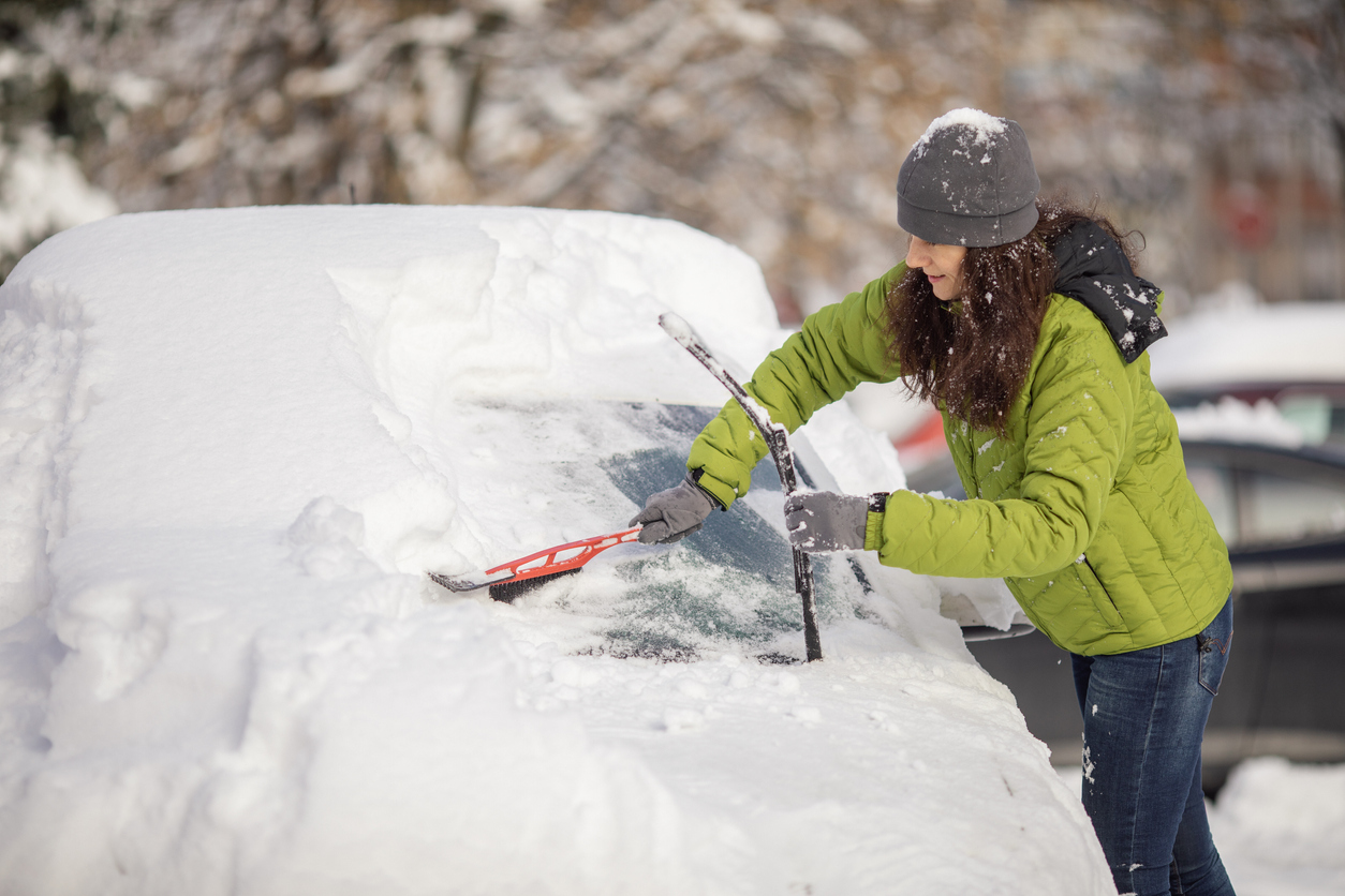 Mature aged woman cleaning her car from snow on parking. She wears warm clothes and using brush for snow