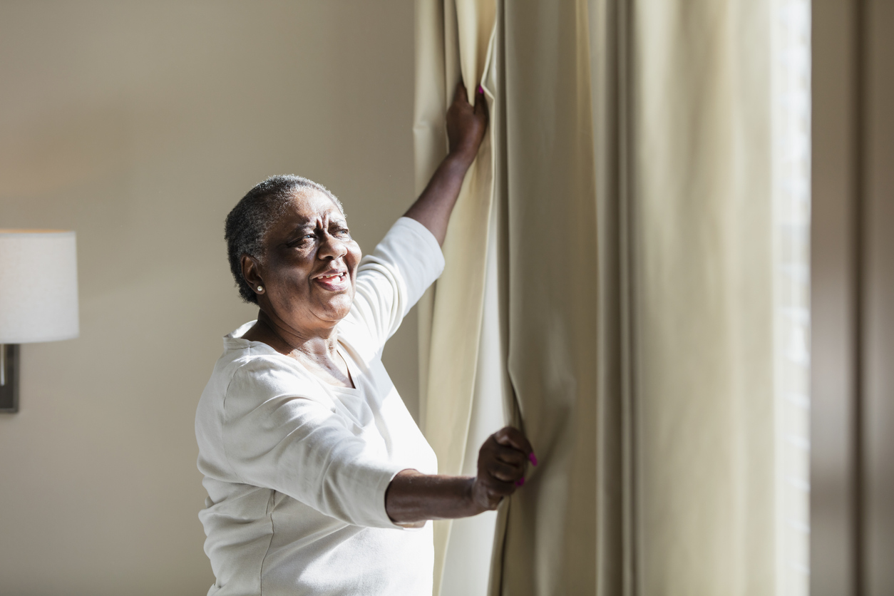 A senior African-American woman in her 70s at home at a window in her bedroom, opening the curtains to let the sunlight in. She has a hopeful expression on her face.
