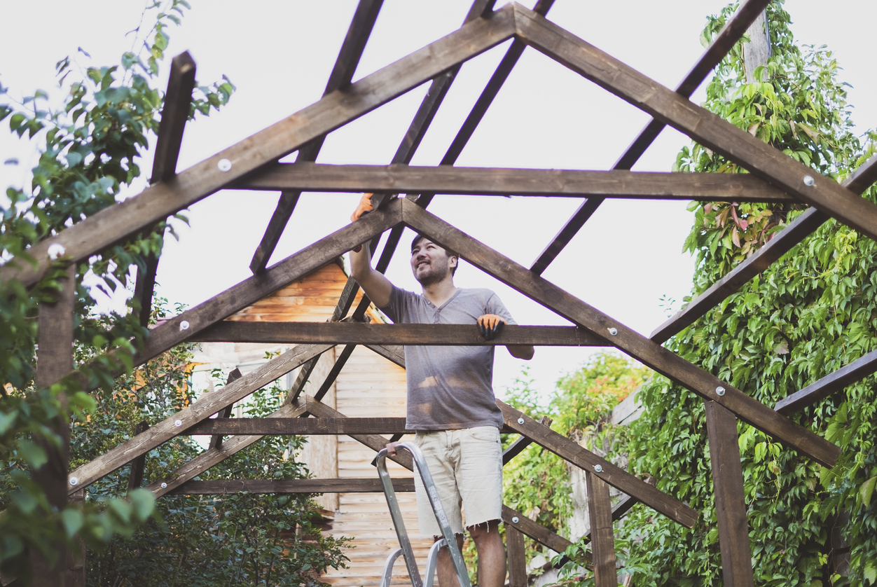 Man with khaki shorts builds pergola structure in green backyard.